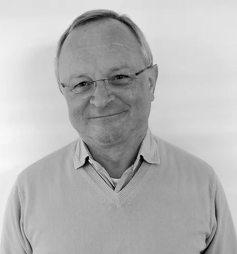 Ein Mann steht vor einem weißen Hintergrund. Er trägt einen Pullover mit V-Ausschnitt über einem Hemd und lächelt leicht in die Kamera.