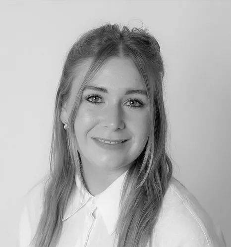 Schwarz-weiß Porträt einer lächelnden Person mit langen Haaren, die ein Hemd mit Kragen trägt. Hintergrund neutral. Ausdruck freundlich und ruhig.