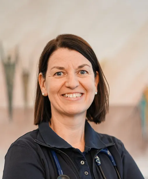 A female medical professional with short hair, wearing a stethoscope and dark uniform, stands smiling in a well-lit room with soft background colors.