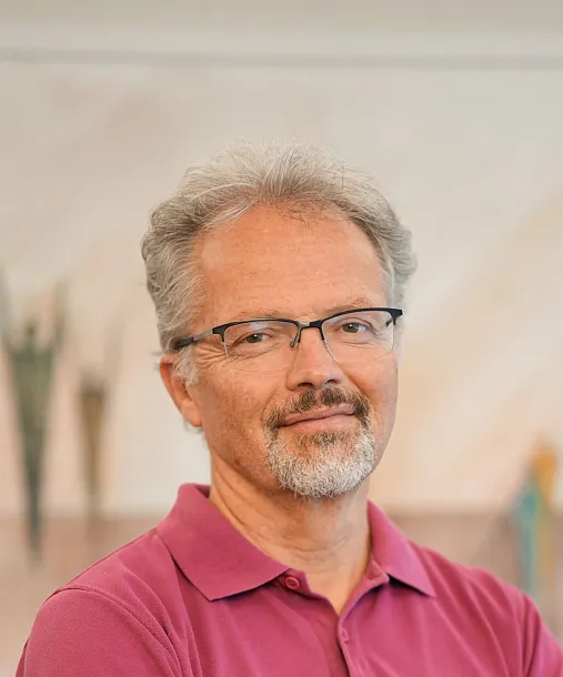 Ein Mann mit grauen Haaren und Brille trägt ein weinrotes Polohemd. Er steht mit verschränkten Armen vor einem verschwommenen Hintergrund in Innenräumen.