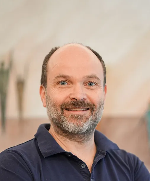 A person with a beard and short hair smiles, wearing a dark blue polo shirt. The background shows abstract wall decorations.