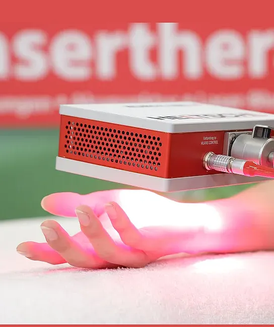 Eine Hand wird von einem Gerät mit rotem Licht bestrahlt, vermutlich für eine Lasertherapie. Der Hintergrund ist unscharf und in warmen Farbtönen gehalten.
