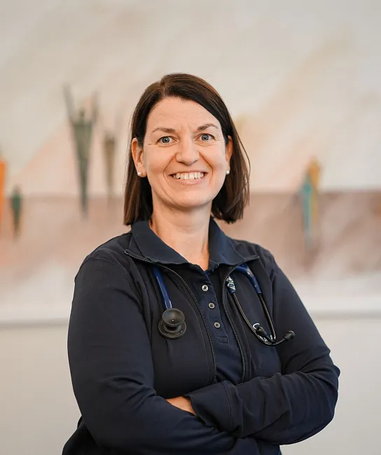 Eine lächelnde Frau in einem medizinischen Outfit mit Stethoskop um den Hals steht vor einem abstrakten Kunstwerk. Sie hat dunkles Haar und trägt Ohrringe.