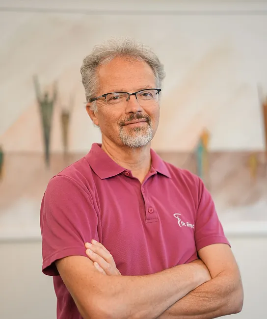Ein Mann mit grauen Haaren und Brille trägt ein weinrotes Polohemd. Er steht mit verschränkten Armen vor einem verschwommenen Hintergrund in Innenräumen.