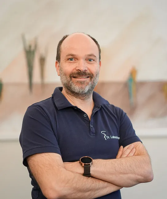 Ein Mann mit Bart lächelt in die Kamera. Er trägt ein dunkelblaues Polohemd mit einem Logo und steht vor einem unscharfen Hintergrund.